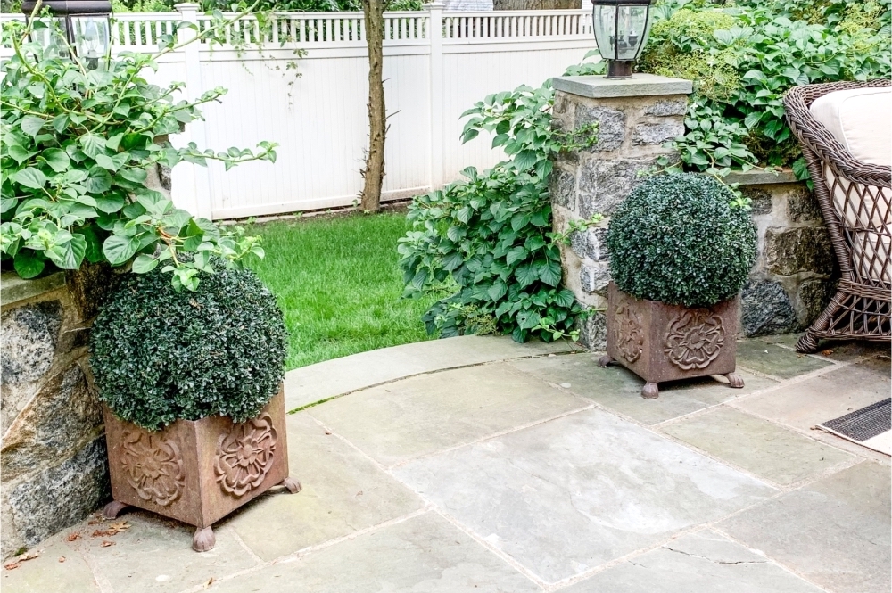 A pair of cast iron garden planters, each on four supports filled with boxwood adorn a patio entryway. The landscaping just beyond features natural plantings and green grass.